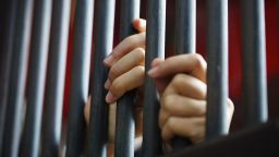 The image is a close-up of two hands wrapped around the bars of a prison cell.
