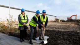 Woman and two men in hi vis jackets and hard hats with a spade digging into the ground, digger, blue sky and a very plain single storey building in background