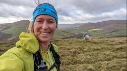 A woman wears a blue headband, a running backpack and a lime green jacket. She holds the camera for a selfie, while behind her are rolling hills and a sheep.