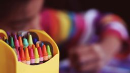 Close up image of crayons with a young person drawing in the background 