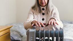 An older woman sits on a bed, holding her hands over a small electric heater.