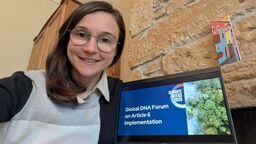 Issi Steeley sat in front of a laptop with the title 'Global DNA Forum on Article 6 Implementation' displayed on the screen