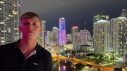 A young male stands in the foreground on a balcony and look towards the camera, resting his arm on the side of the balcony. It is night time and behind them is a brightly light cityscape with tall buildings.