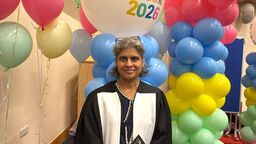A person smiling at the camera, dressed in a black robe, holding a scroll, surrounded by balloons.