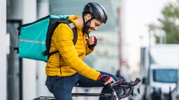 A man on a bike with a food delivery bag on his back, wearing a yellow jacket and helment.