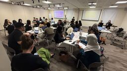 People sat around tables, using laptops and looking at people presenting at the front of the room